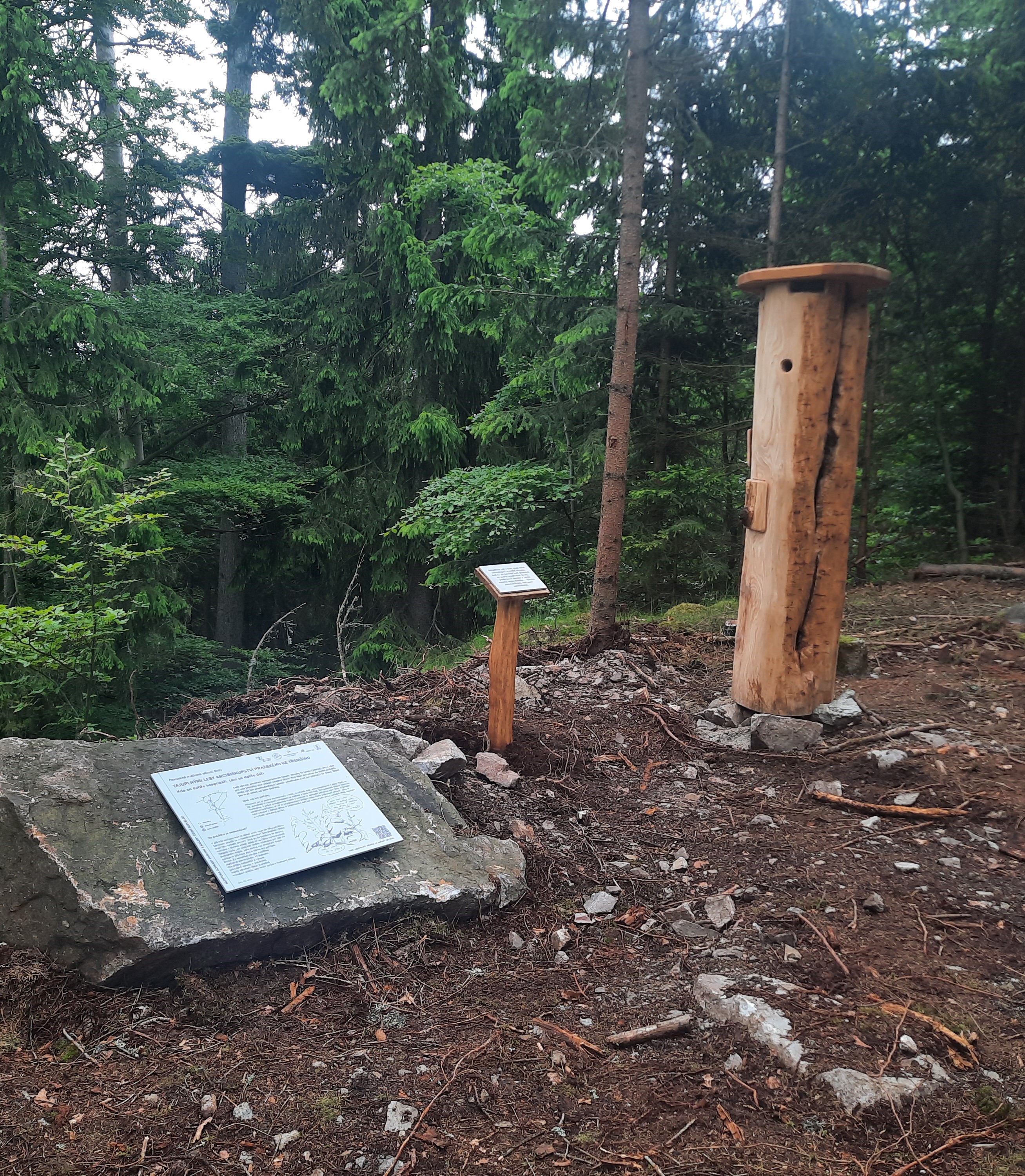 Zastavení naučné stezky s kovovou naučnou tabulí na kameni a interaktivním prvkem v podobě dutého kmene.